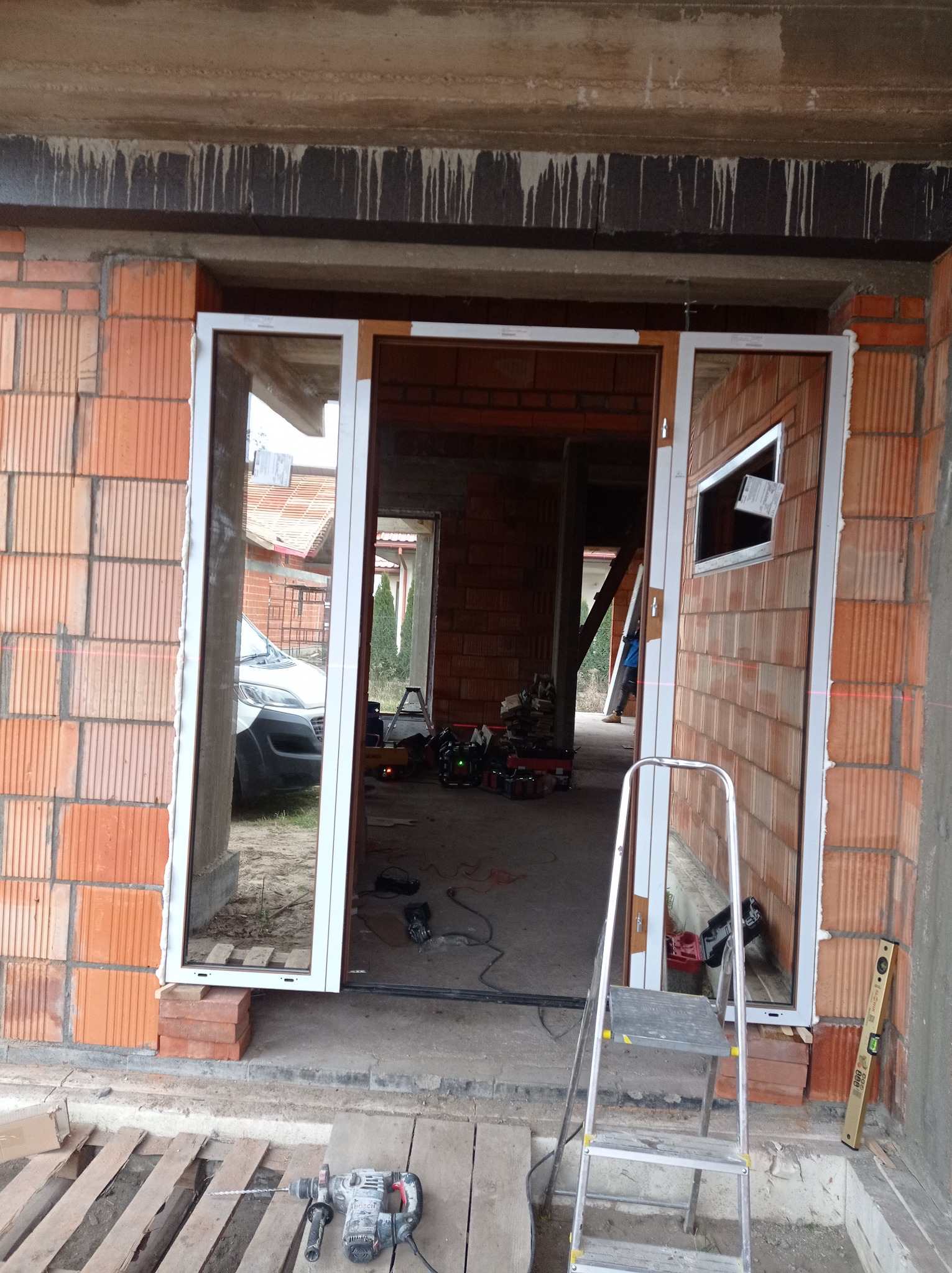 Montaż okna w surowym budynku z czerwonej cegły, widoczna drabina, wiertarka udarowa i poziomica, laserowy wskaźnik poziomu, na zewnątrz zaparkowany samochód dostawczy.