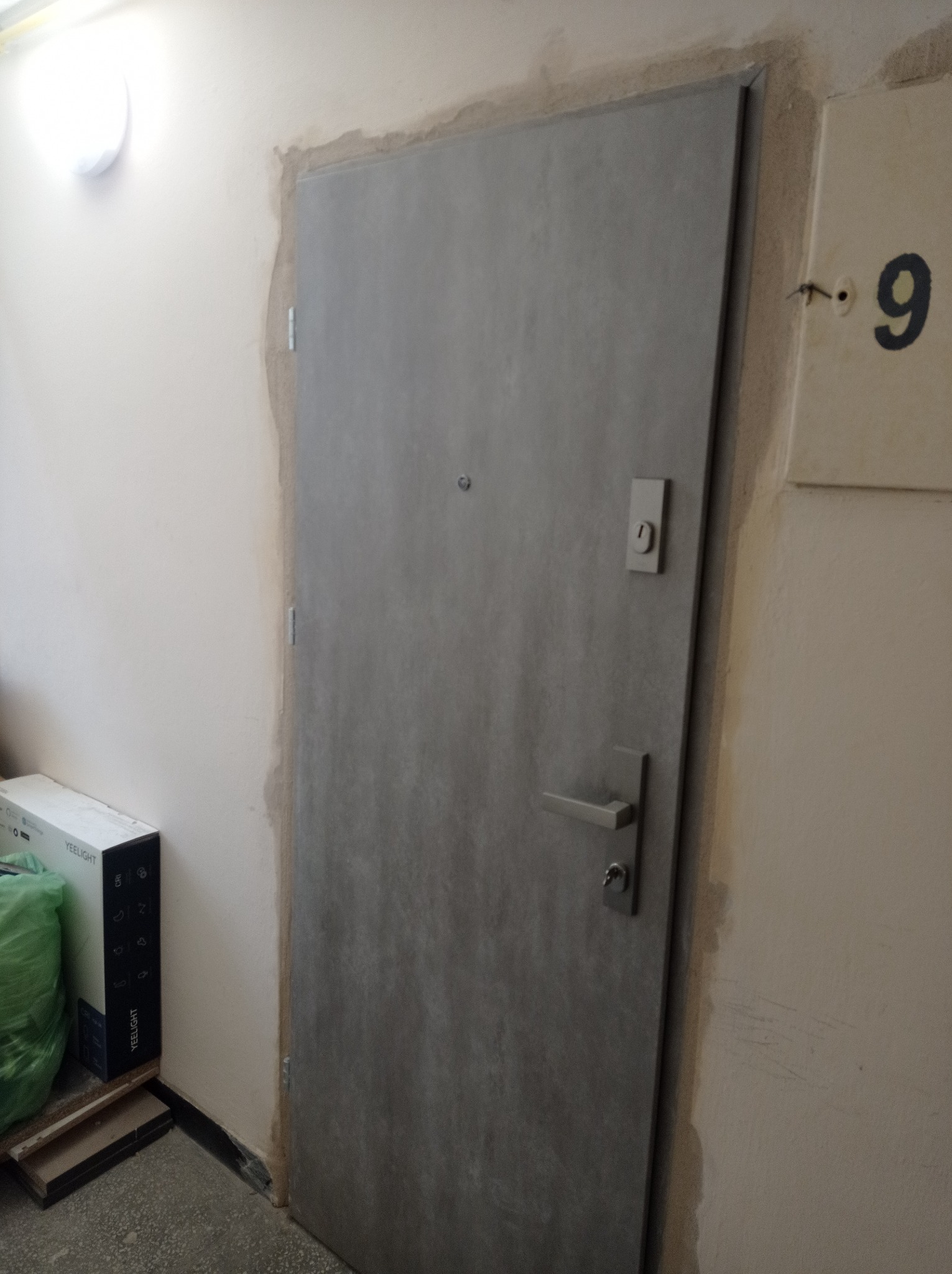 Szare drzwi wejściowe z wizjerem i numerem mieszkania 9 na ścianie, widoczny karton po żarówkach Yeelight, ściana po remoncie.