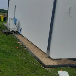 Przygotowanie podłoża pod trawnik z rolki przy ścianie domu z widoczną jednostką zewnętrzną klimatyzacji i rurą spustową.