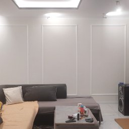 Usługi Wielobranżowe - Salon z narożną sofą, telewizorem z widocznym programem informacyjnym i piecem typu koza z rurą odprowadzającą dym, białe ściany zdobione sztukaterią.