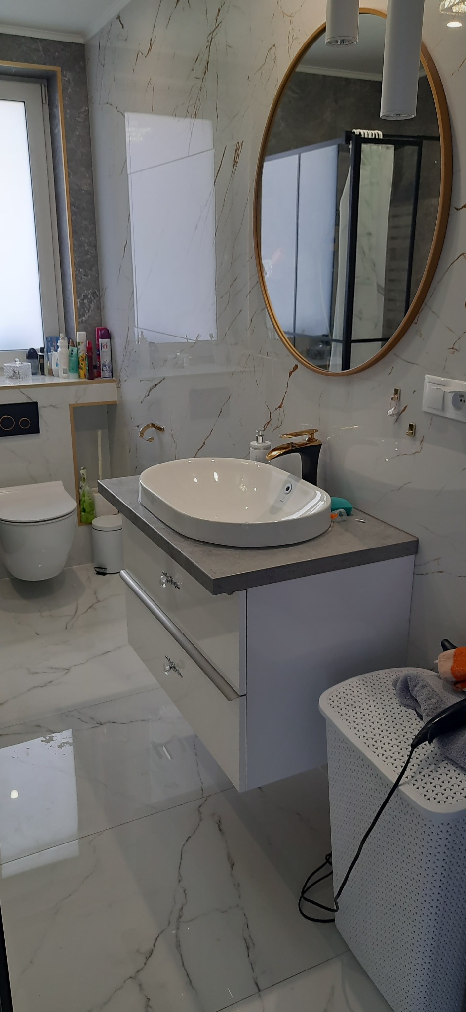 Nowoczesna łazienka z marmurowymi ścianami i podłogą, owalna umywalka nablatowa z baterią w kolorze szczotkowanego złota, okrągłe lustro z podświetleniem LED i szafka pod umywalką z kryształowymi...