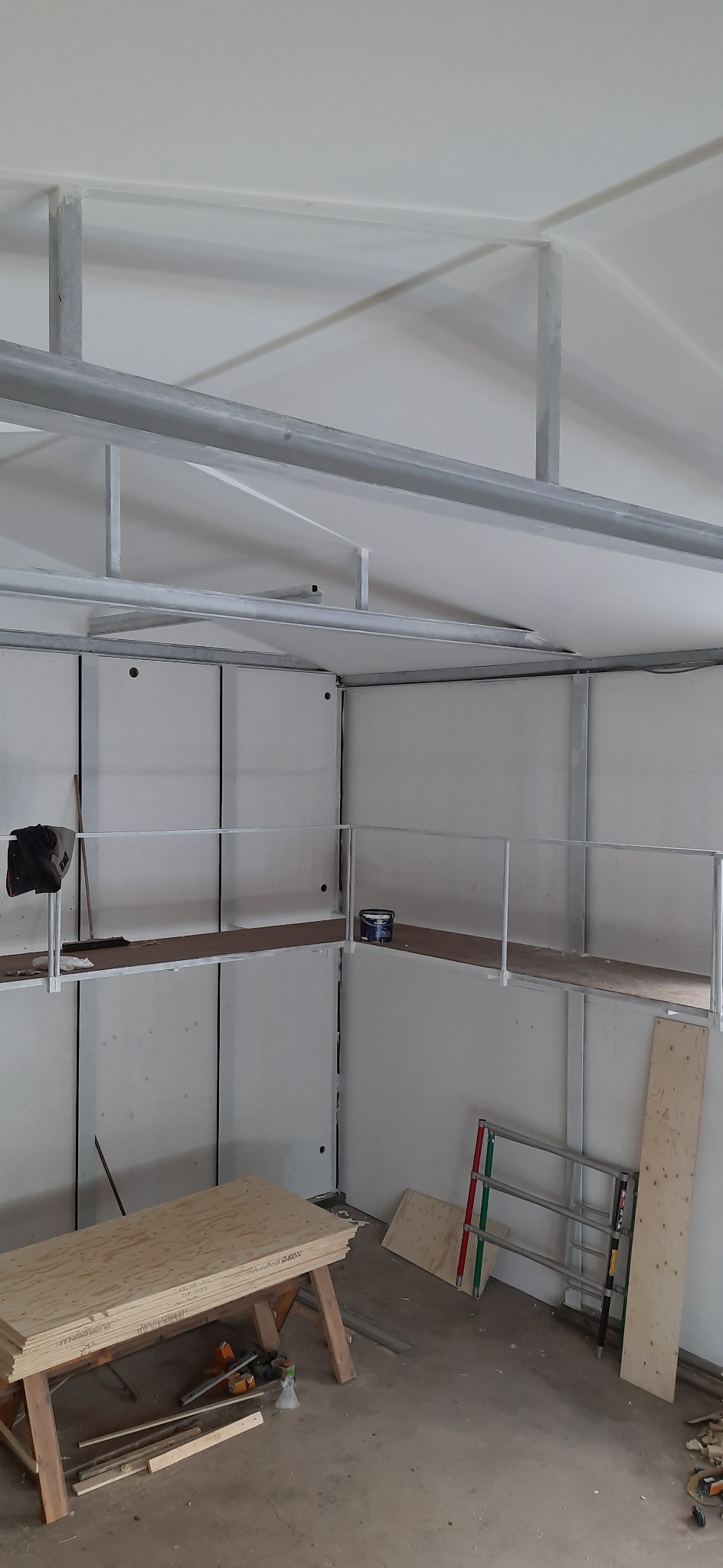 Wnętrze w trakcie budowy z widoczną konstrukcją szkieletową, płytami ściennymi, rusztowaniem, stosem sklejki na kozłach i puszką farby na podwyższeniu.