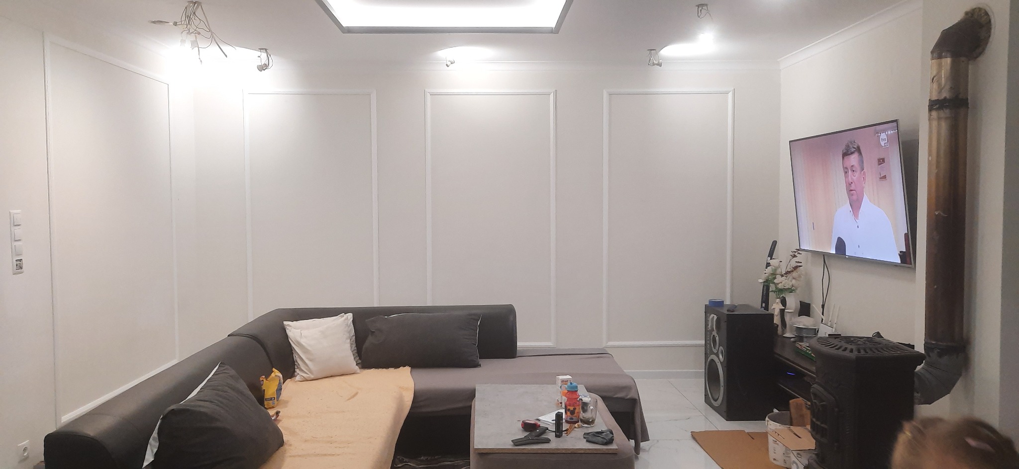 Salon z narożną sofą, telewizorem z widocznym programem informacyjnym i piecem typu koza z rurą odprowadzającą dym, białe ściany zdobione sztukaterią.