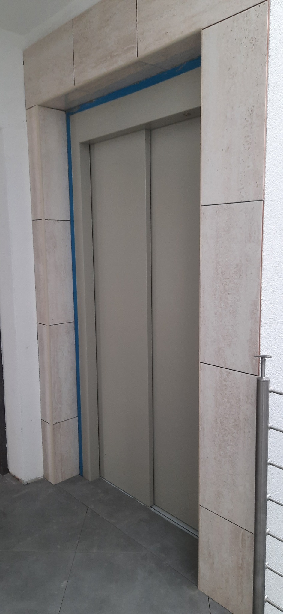 Wykończenie wejścia do windy płytkami z naturalnego kamienia w odcieniach beżu i szarości, zabezpieczone niebieską taśmą malarską.