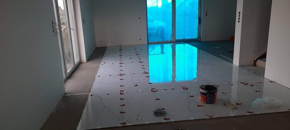 Wnętrze w trakcie układania błyszczących, białych płytek podłogowych z szarym wzorem żyłek, widoczne czerwone krzyżyki dystansowe, wiadro z klejem i okna zabezpieczone folią.