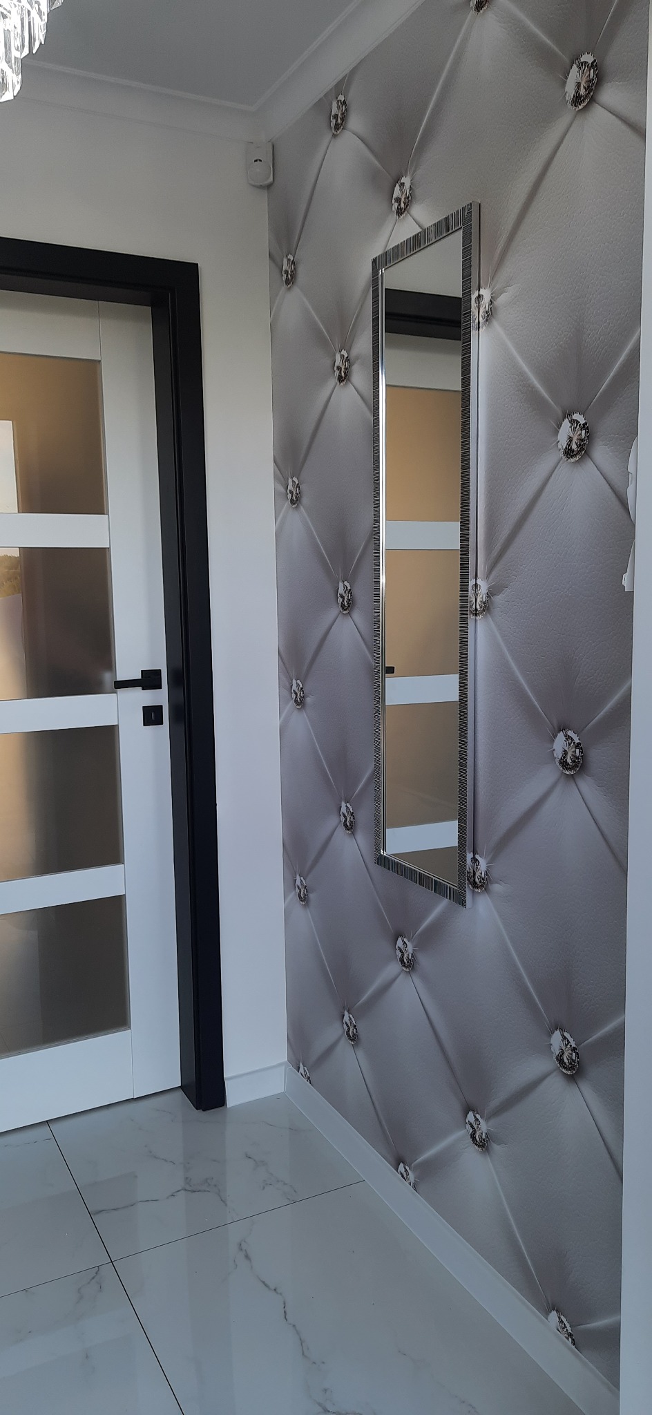 Fragment korytarza z białą, przeszkloną ścianką działową, tapetą imitującą pikowaną skórę z kryształkami i wąskim lustrem na ścianie oraz białą podłogą z marmurowym wzorem.