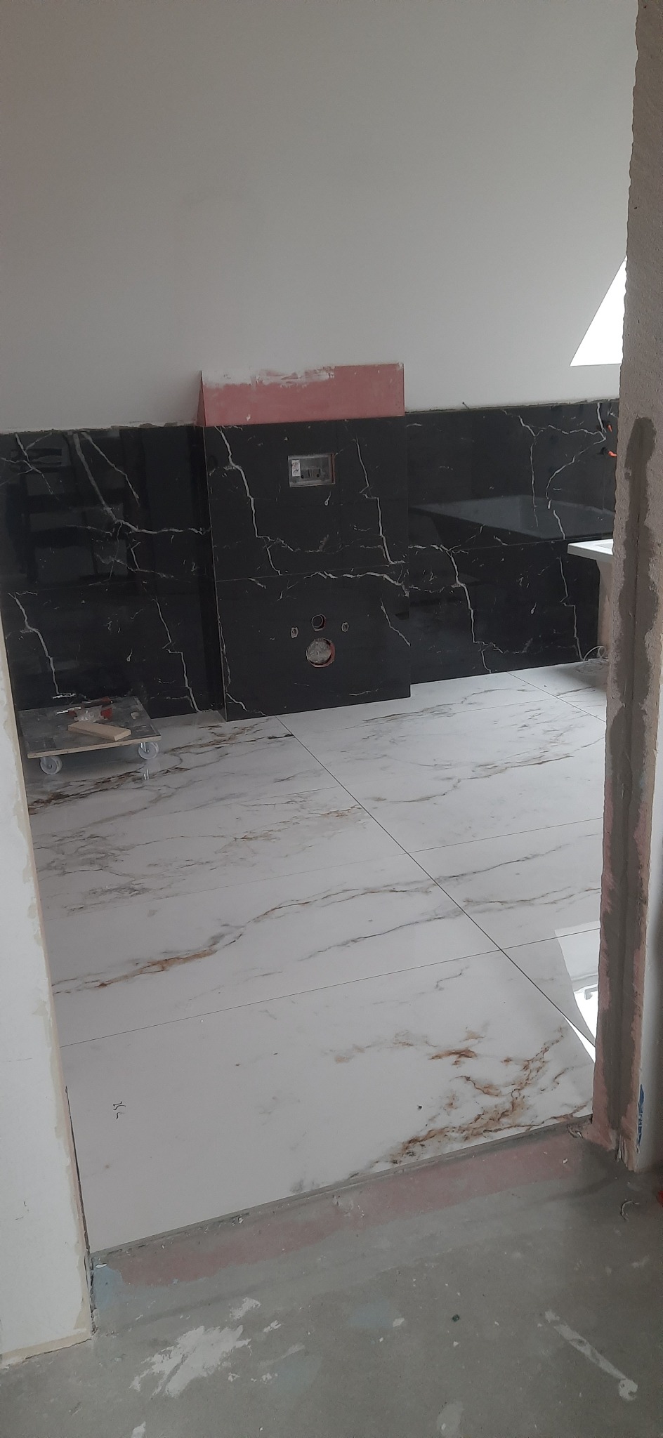 Częściowo wyremontowana łazienka z czarnymi płytkami na ścianach z białymi żyłami i białymi płytkami na podłodze z brązowymi żyłami, widoczne przygotowania do montażu armatury oraz wózek...