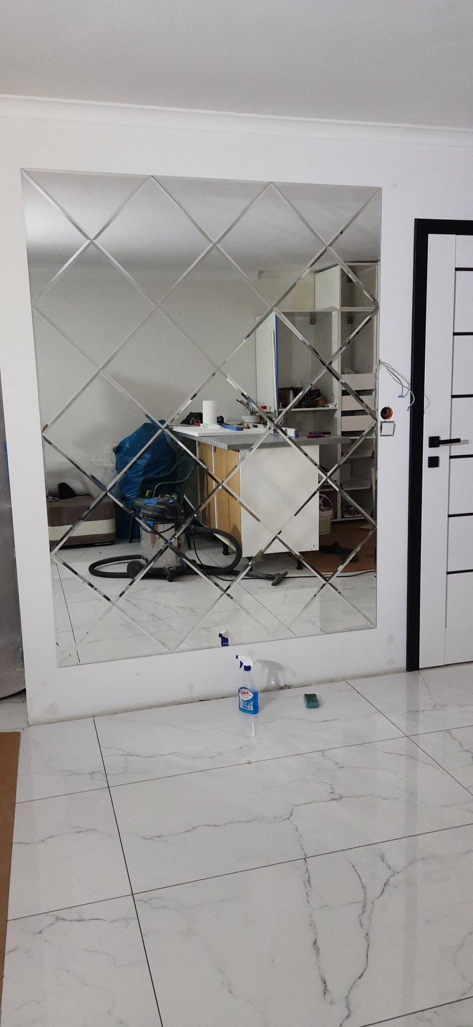 Ściana wyłożona lustrzanymi rombami w łazience, widoczny odkurzacz i środki czystości na podłodze z marmurowych płytek.