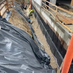 OLTA SP ZOO - Budowa fundamentów budynku z izolacją przeciwwilgociową i warstwą drenażową z otoczaków, widoczni pracownicy w żółtych kamizelkach i czarnych rur drenażowych.