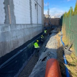 OLTA SP ZOO - Budowa drenażu: robotnicy w żółtych kamizelkach układają rury i folię izolacyjną w wykopie przy fundamencie budynku z białego bloczka.