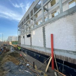 OLTA SP ZOO - Budowa budynku w trakcie, widoczny fundament zabezpieczony folią i żwirem, robotnicy w odblaskowych kamizelkach przy pracy, pomarańczowa rura pionowo przy ścianie, niebo z chmurami.