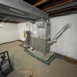 OLTA SP ZOO - Instalacja systemu odprowadzania wody w piwnicy, z widocznym bojlerem i piecem, otoczonymi zielonymi rurkami drenażowymi i kamieniami, w surowym wnętrzu z betonową podłogą i odsłoniętą konstrukcją...