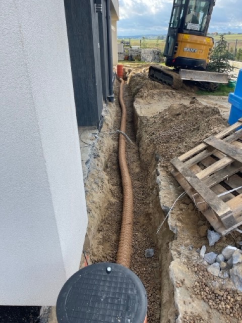 Brązowa perforowana rura ułożona w wykopie wzdłuż ściany budynku, z widocznym żółtym minikoparką w tle i paletą obok wykopu.