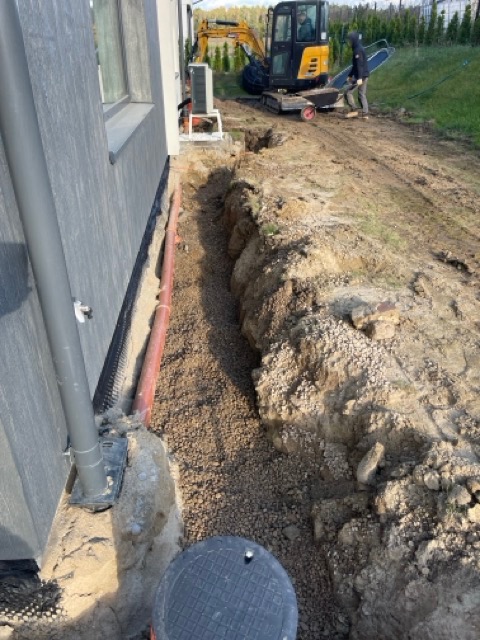 Prace ziemne przy budynku w Warszawie: wykop z rurą i drenażem obsypany żwirem, w tle koparka i pracownik.