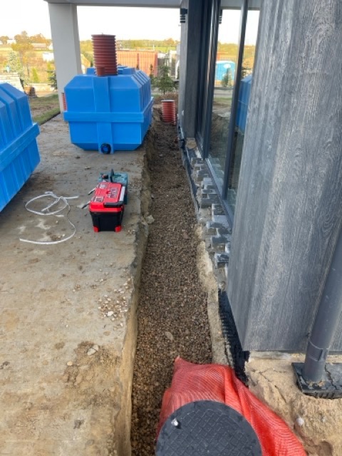 Wykop wzdłuż fundamentu domu wypełniony żwirem, widoczne niebieskie zbiorniki retencyjne i skrzynka z narzędziami, sugerujące prace budowlane lub instalacyjne.