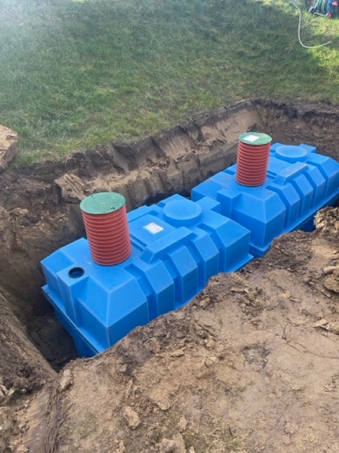 Montaż dwóch, niebieskich zbiorników retencyjnych w wykopie ziemnym, z widocznymi elementami przyłączeniowymi w kolorze czerwono-zielonym.