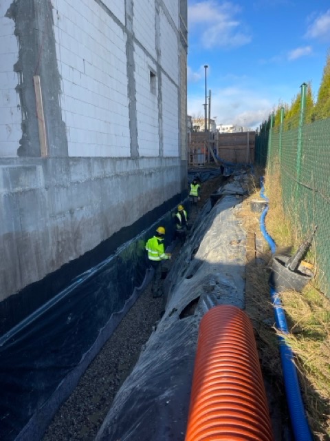 Budowa drenażu: robotnicy w żółtych kamizelkach układają rury i folię izolacyjną w wykopie przy fundamencie budynku z białego bloczka.