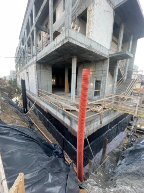 Budynek w stanie surowym otwartym z widoczną izolacją fundamentów i pomarańczową rurą drenarską na pierwszym planie, czarna folia zabezpieczająca wykop.