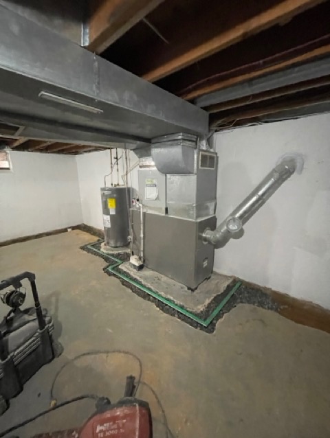 Instalacja systemu odprowadzania wody w piwnicy, z widocznym bojlerem i piecem, otoczonymi zielonymi rurkami drenażowymi i kamieniami, w surowym wnętrzu z betonową podłogą i odsłoniętą konstrukcją...