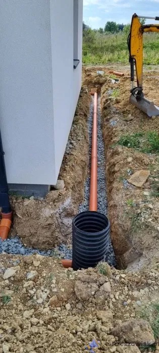 Pomarańczowa rura drenażowa ułożona w wykopie wypełnionym żwirem, obok budynku, z czarną rurą karbowaną w dole i koparką w tle.