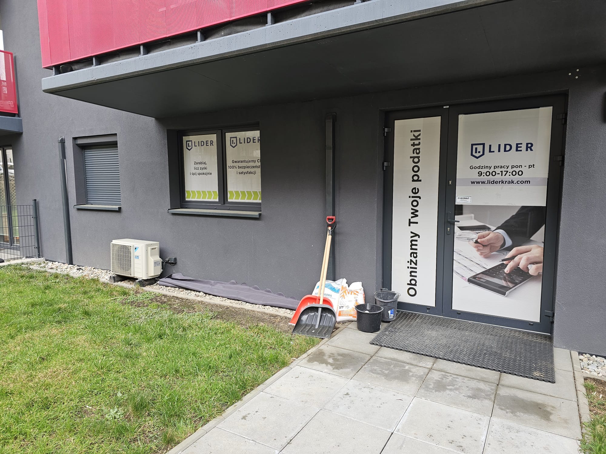 Wejście do biura firmy Lider z widocznymi oklejonymi szybami z informacjami o ofercie, obok ściany oparta łopata i worki z solą, przed budynkiem trawnik.