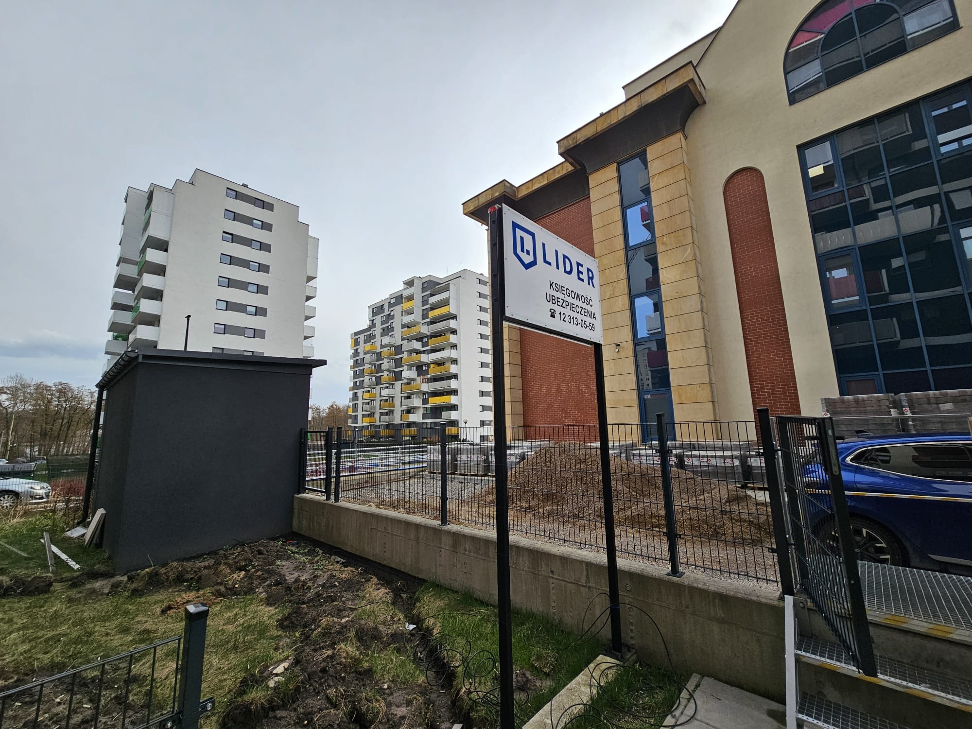 Tablica informacyjna z logo 'LIDER Księgowość Ubezpieczenia' na tle budynków mieszkalnych i biurowca z ceglanymi elementami.