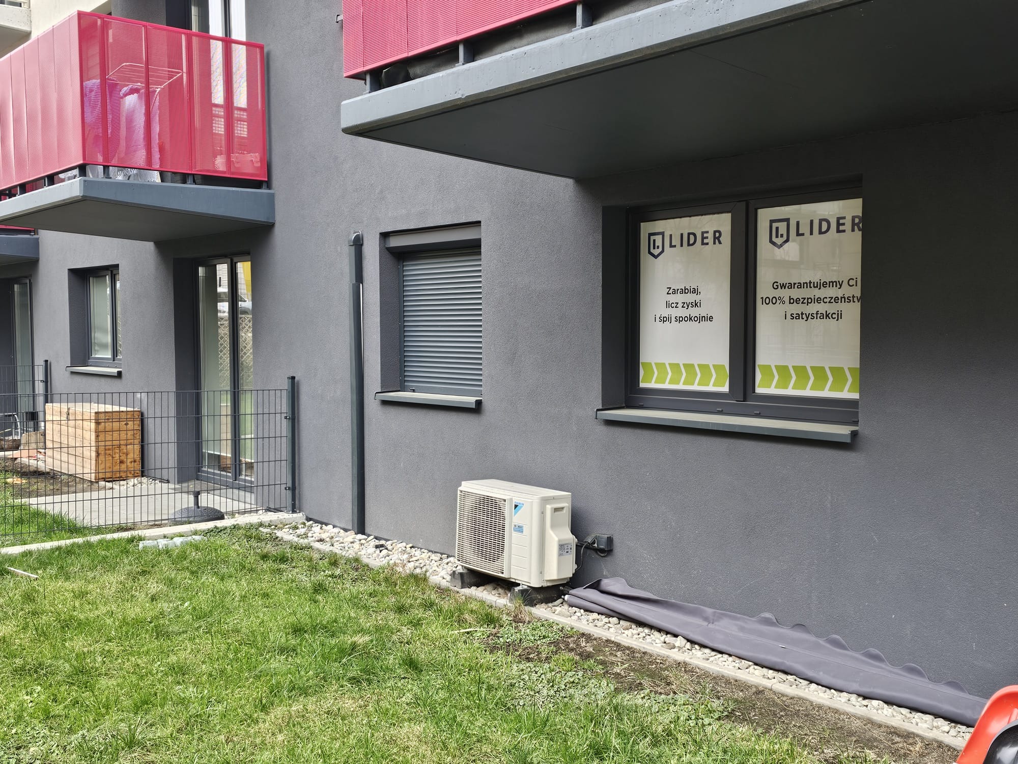 Zewnętrzna jednostka klimatyzacji Daikin przy szarym budynku z czerwonymi balkonami i oknami z naklejkami reklamowymi Lider, widok z trawnika.