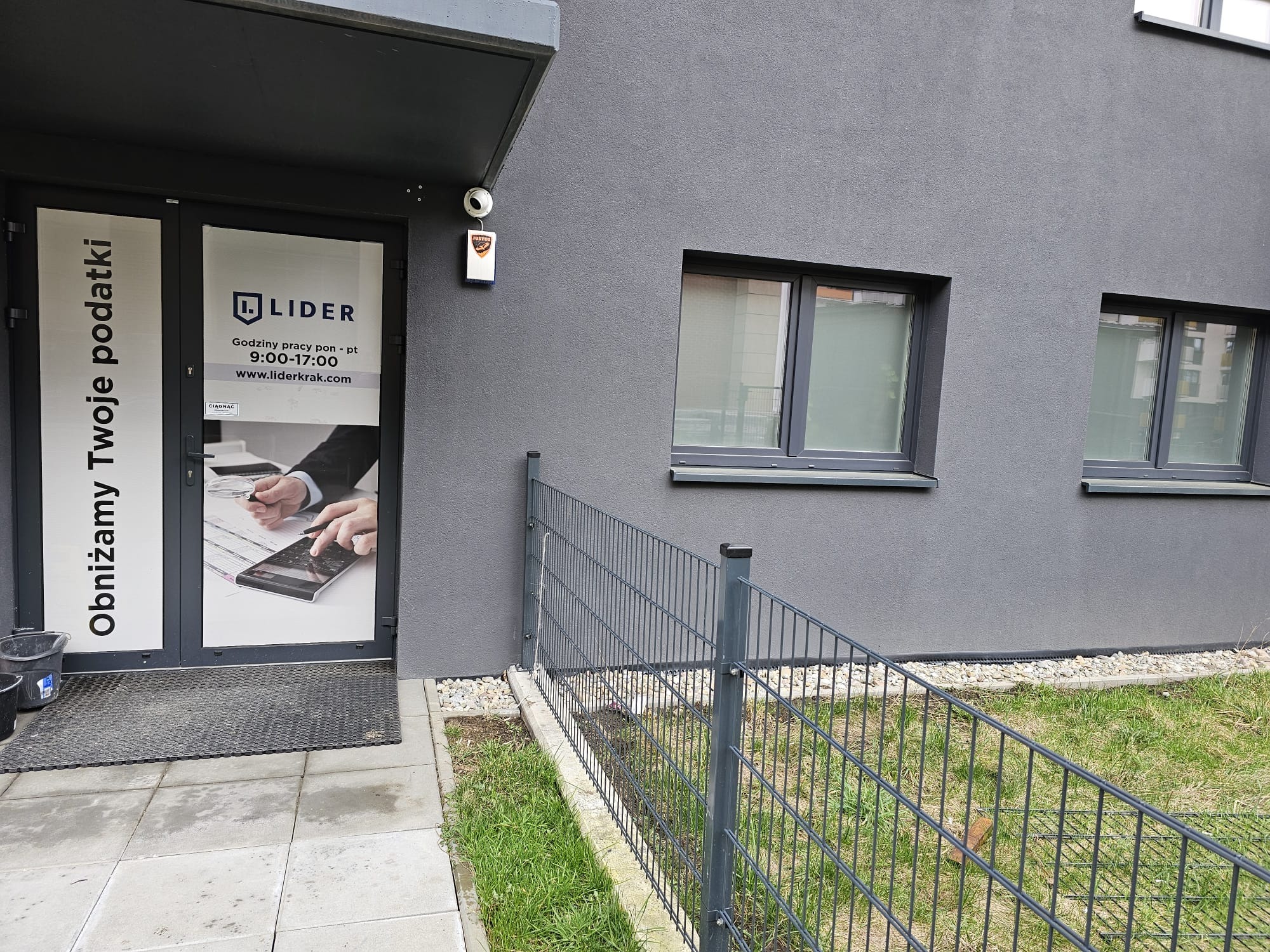 Wejście do biura firmy Lider, oferującej usługi obniżania podatków, z grafiką przedstawiającą ręce analizujące dokumenty i kalkulator, widoczne godziny pracy i adres strony internetowej na drzwiach.