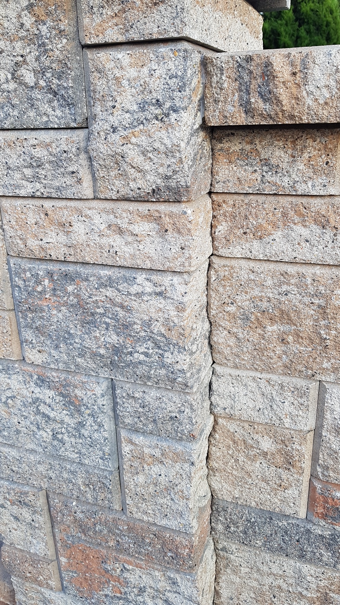 Fragment muru z bloków betonowych o nieregularnej fakturze i zróżnicowanej kolorystyce, widoczne łączenia między elementami, na górze widoczny fragment zieleni.