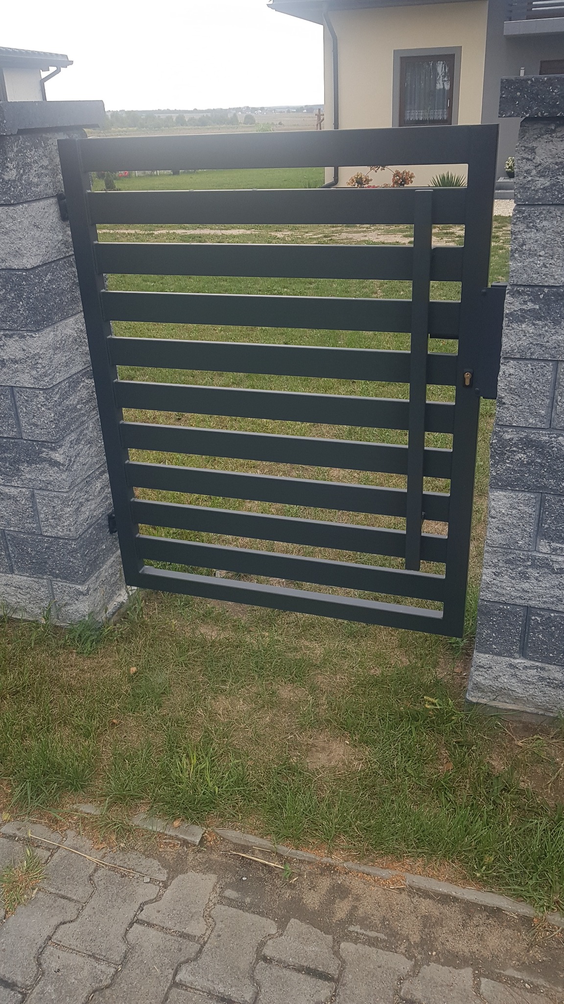 Nowoczesna, pozioma brama wjazdowa w kolorze grafitowym, zamontowana między dwoma słupkami z szarej kostki betonowej, widok na trawnik i dom w tle.