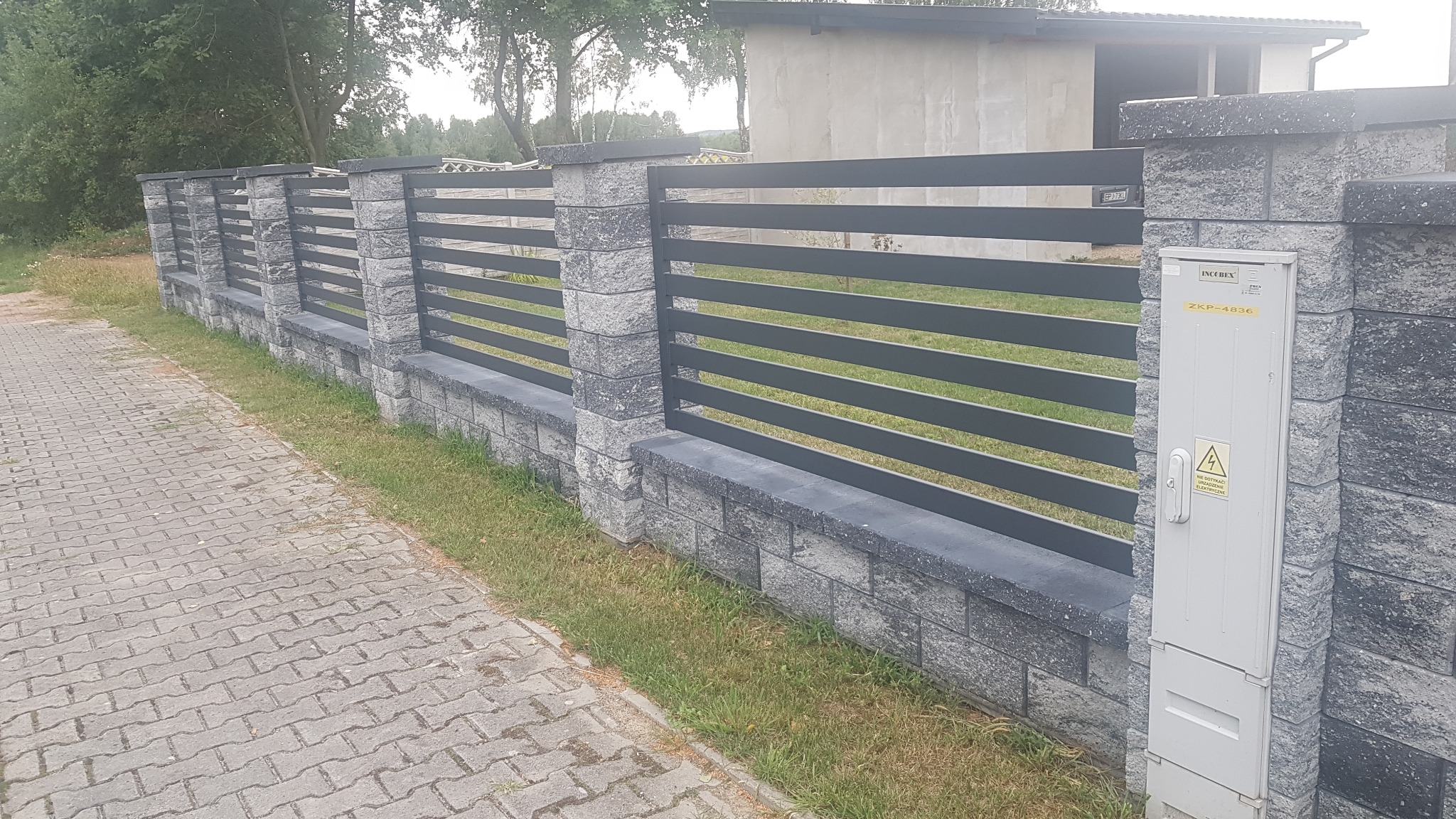 Nowoczesne ogrodzenie z szarych bloczków betonowych i poziomych, metalowych przęseł, z widoczną skrzynką elektryczną na jednym ze słupków, ułożone wzdłuż chodnika z kostki brukowej.