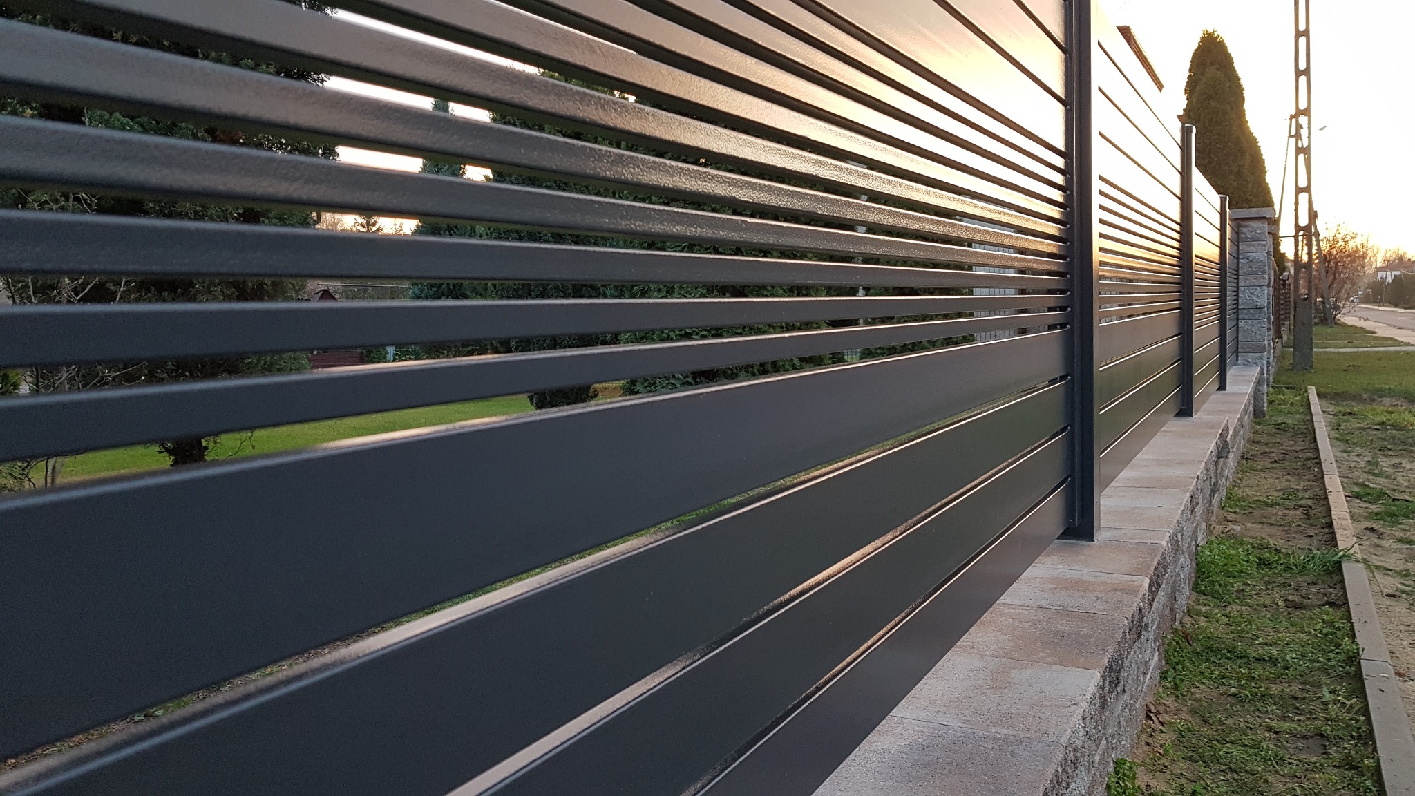 Nowoczesne, poziome przęsła stalowe w kolorze grafitowym, tworzące ogrodzenie na niskiej, kamiennej podmurówce, widok z boku w kierunku zachodzącego słońca.