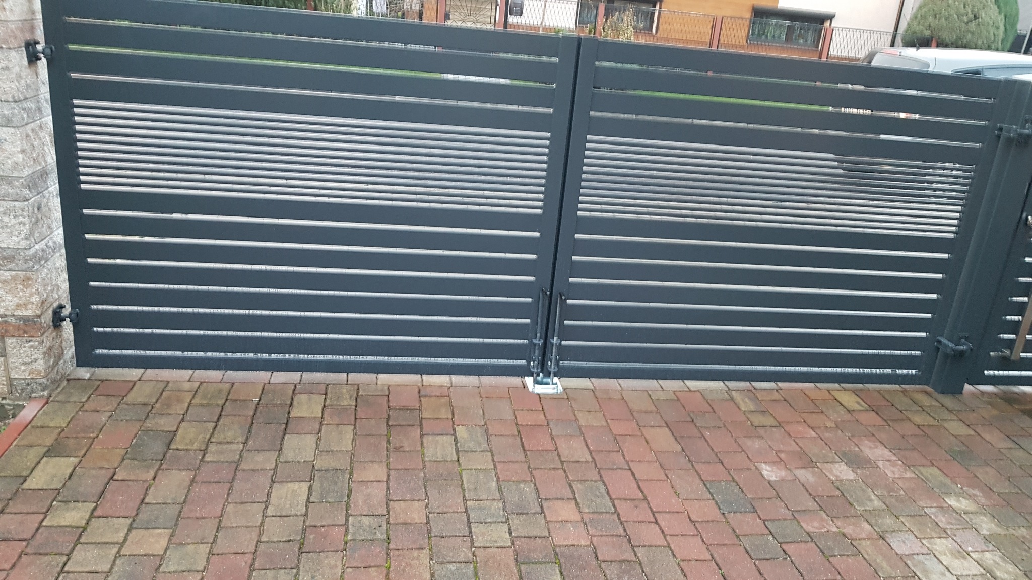 Nowoczesna, pozioma brama wjazdowa z metalowymi lamelami w kolorze grafitowym, zamontowana na kostce brukowej w odcieniach czerwieni i brązu.