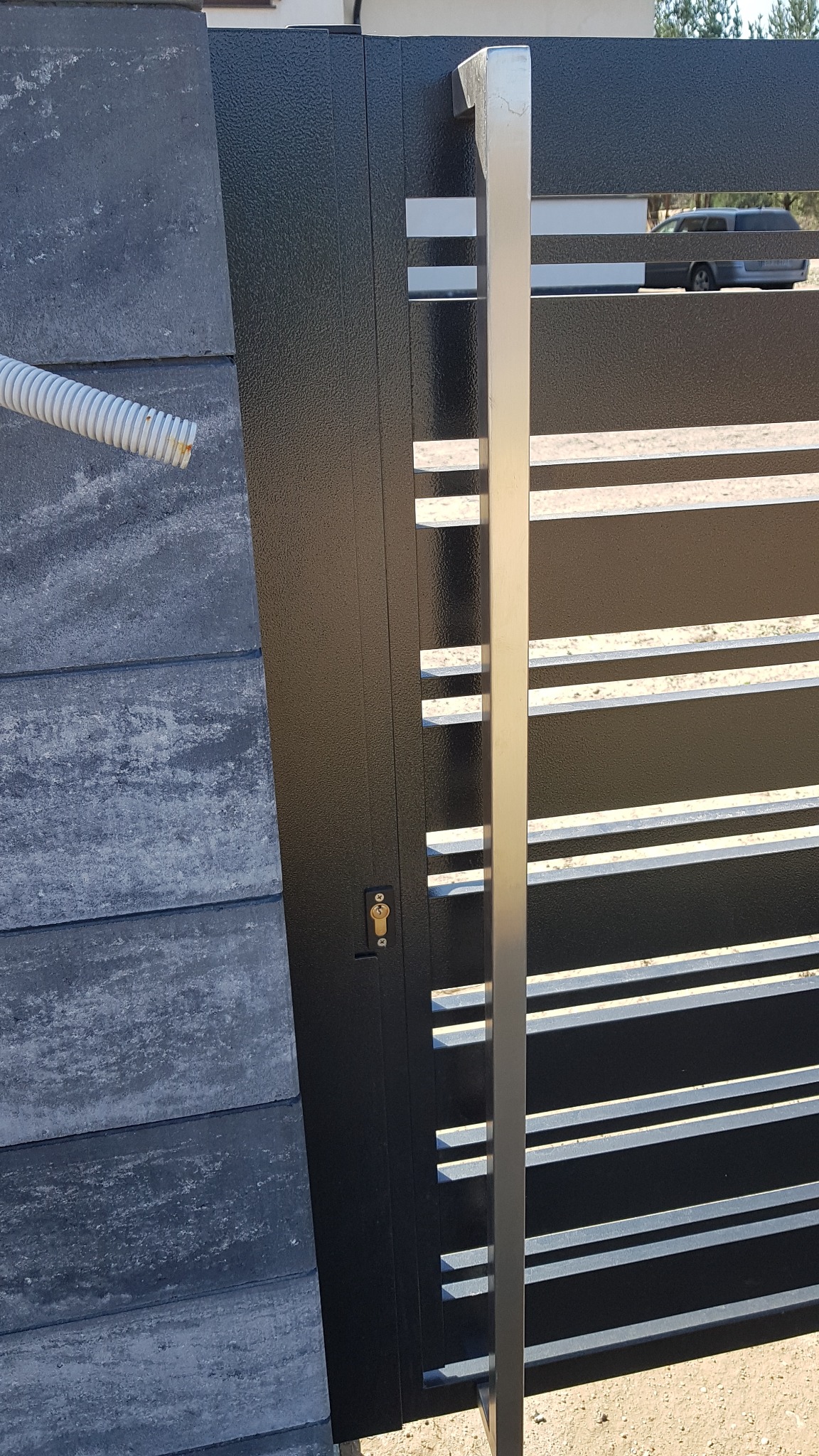 Nowoczesna brama wjazdowa z czarnymi, poziomymi profilami i srebrnym uchwytem, przylegająca do betonowego ogrodzenia, z widocznym kluczem w zamku i fragmentem węża instalacyjnego.