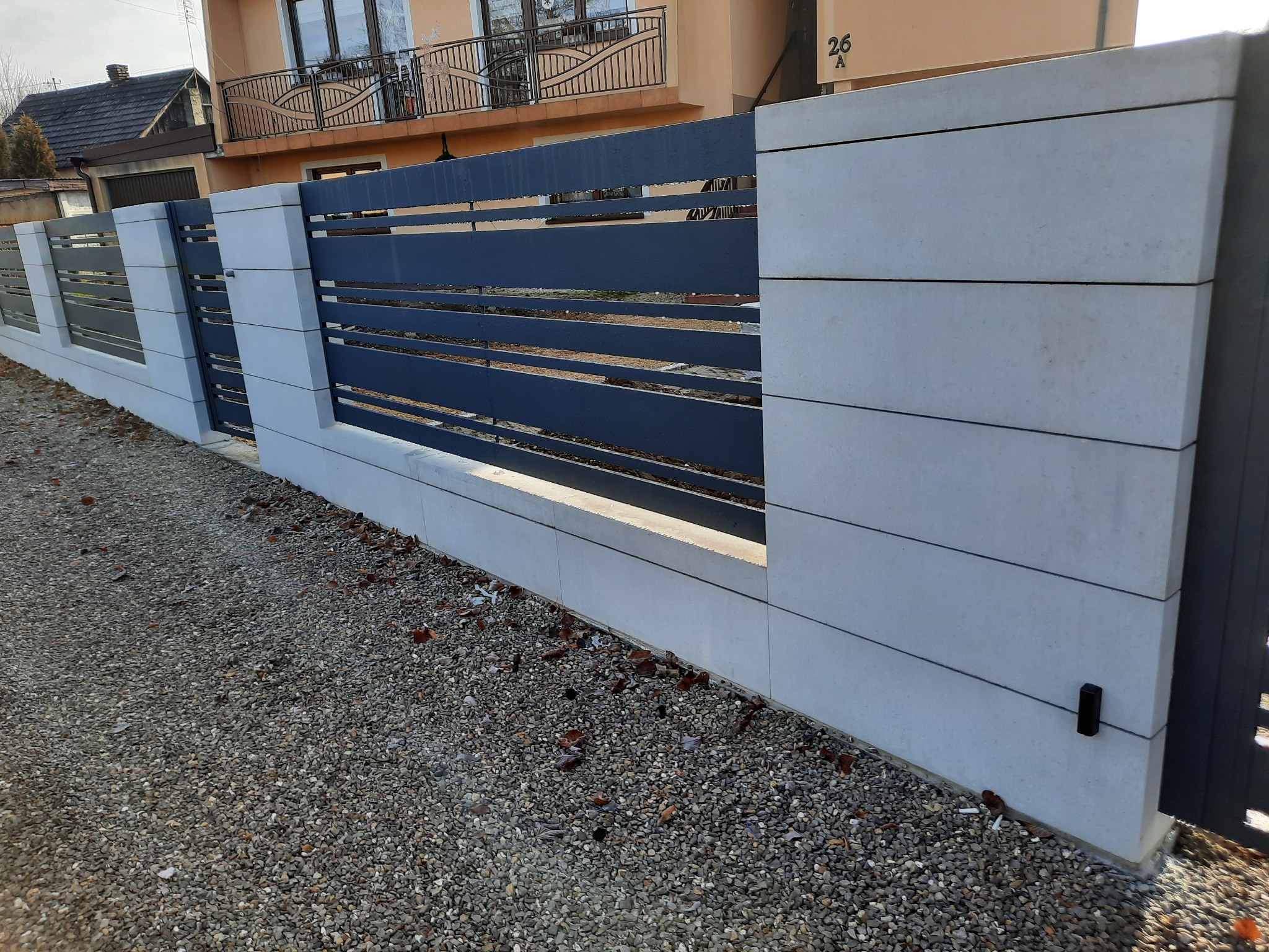 Nowoczesne ogrodzenie z szarych, betonowych segmentów i poziomych, stalowych przęseł w kolorze antracytowym; widok z poziomu gruntu na tle domu z balkonem.