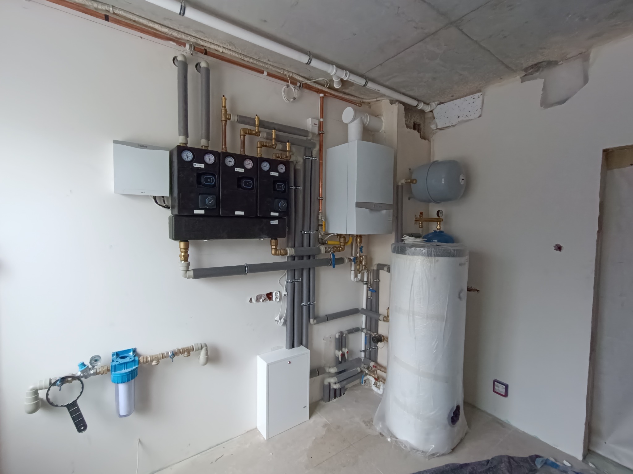 Instalacja kotła grzewczego z osprzętem, manometrami i filtrem wody w surowym wnętrzu z widocznym betonowym stropem.