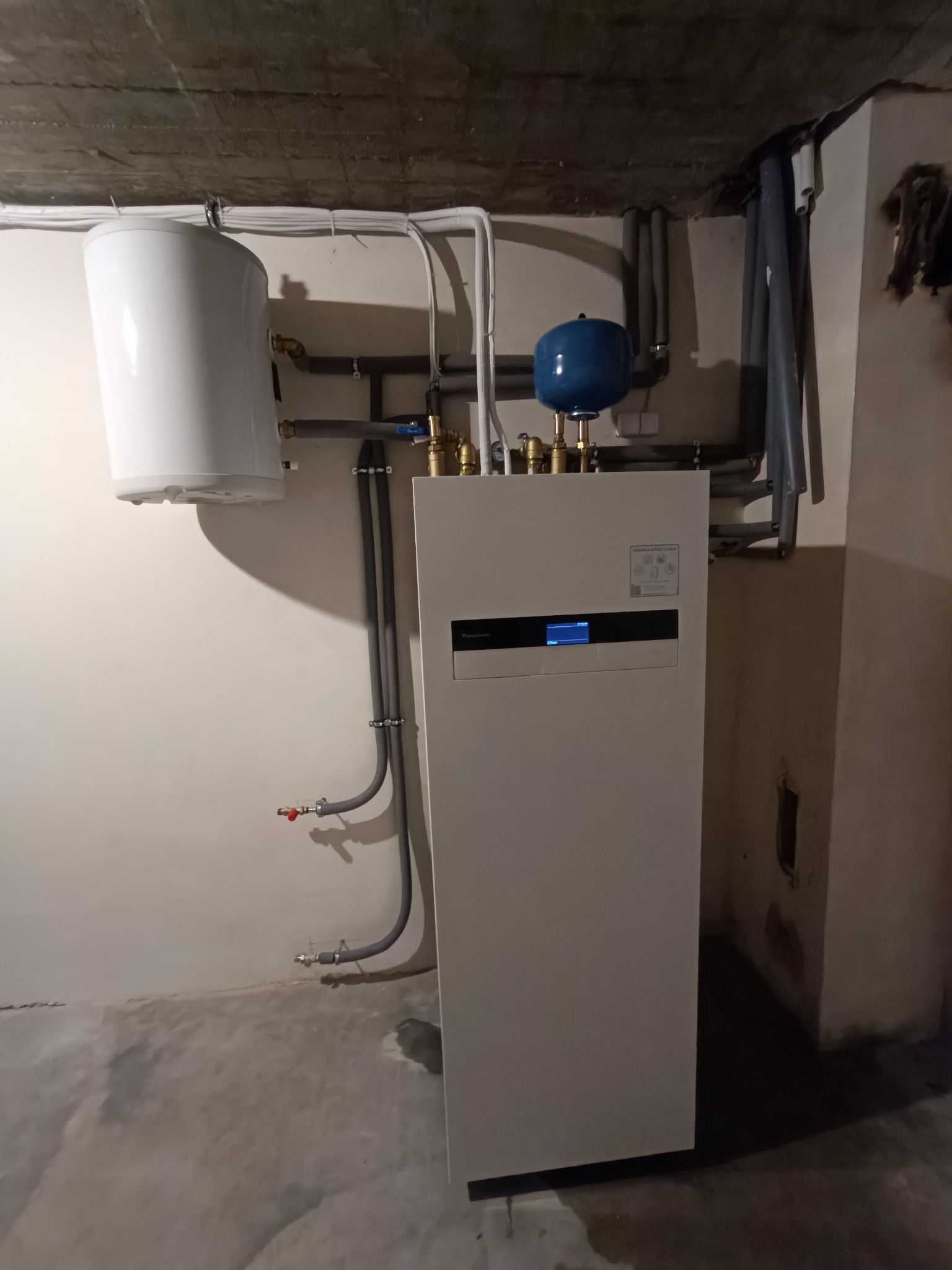 Biały piec grzewczy z niebieskim zbiornikiem wyrównawczym i białym zasobnikiem ciepłej wody, zamontowane w surowym pomieszczeniu z betonowym sufitem i ścianami, widoczne rury instalacyjne.