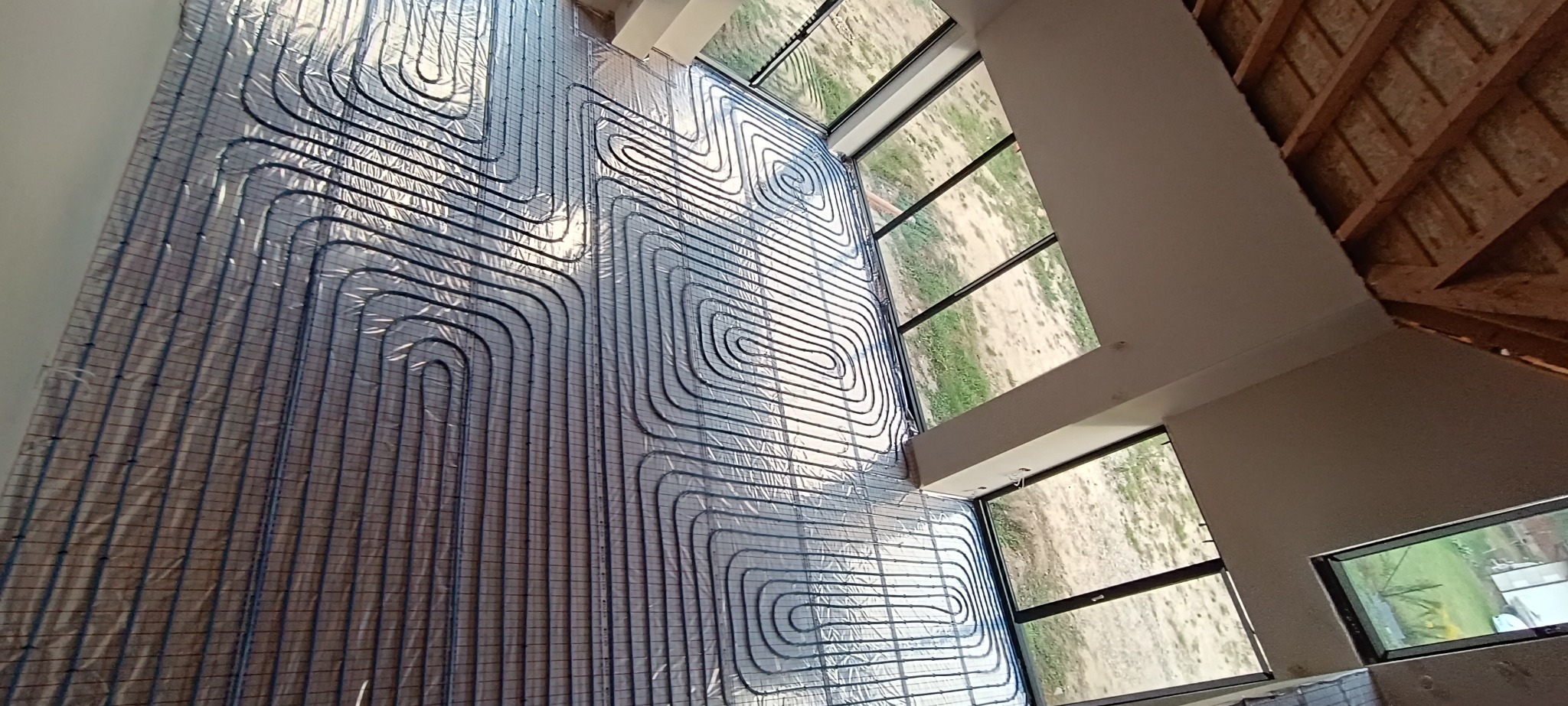 Rozłożona instalacja ogrzewania podłogowego z widocznymi rurami na folii izolacyjnej, duże okna w tle, surowy stan wykończenia.