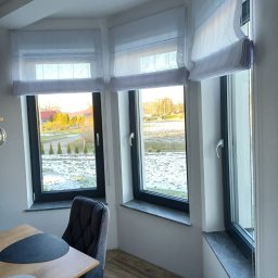 Cortina Mariola Świerk - Trzy okna z zamontowanymi białymi roletami rzymskimi, widok na zaśnieżony krajobraz, w tle budynki i drzewa.