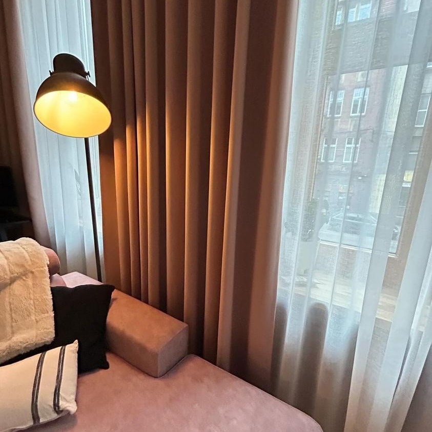 Beżowe zasłony zaciemniające z regularnymi fałdami, obok okna z delikatnymi, białymi firanami i lampą podłogową w rogu pokoju, widok na budynek za oknem.