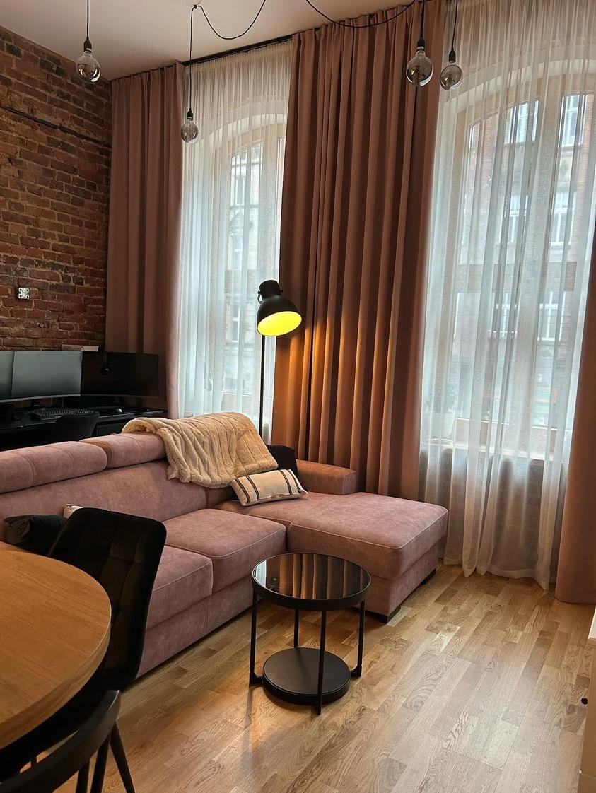 Salon z ceglaną ścianą, różową sofą i wysokimi oknami przesłoniętymi brązowymi zasłonami oraz białymi firanami