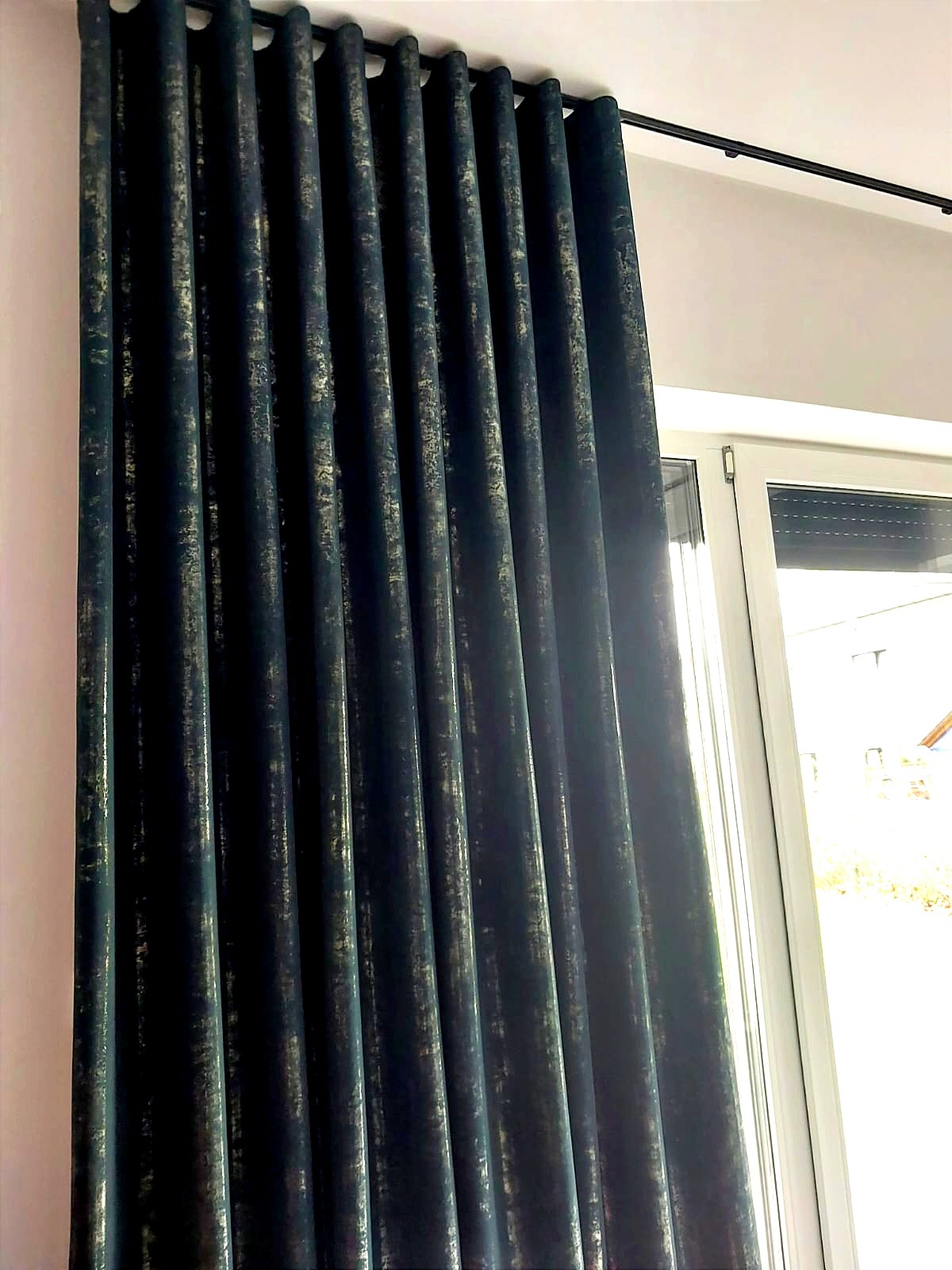 Elegancka, długa, ciemna zasłona z delikatnym, złotym wzorem, zawieszona na czarnym karniszu w Bytomiu. Widoczne okno z częściowo zasłoniętą roletą zewnętrzną. Zasłona tworzy regularne fałdy.