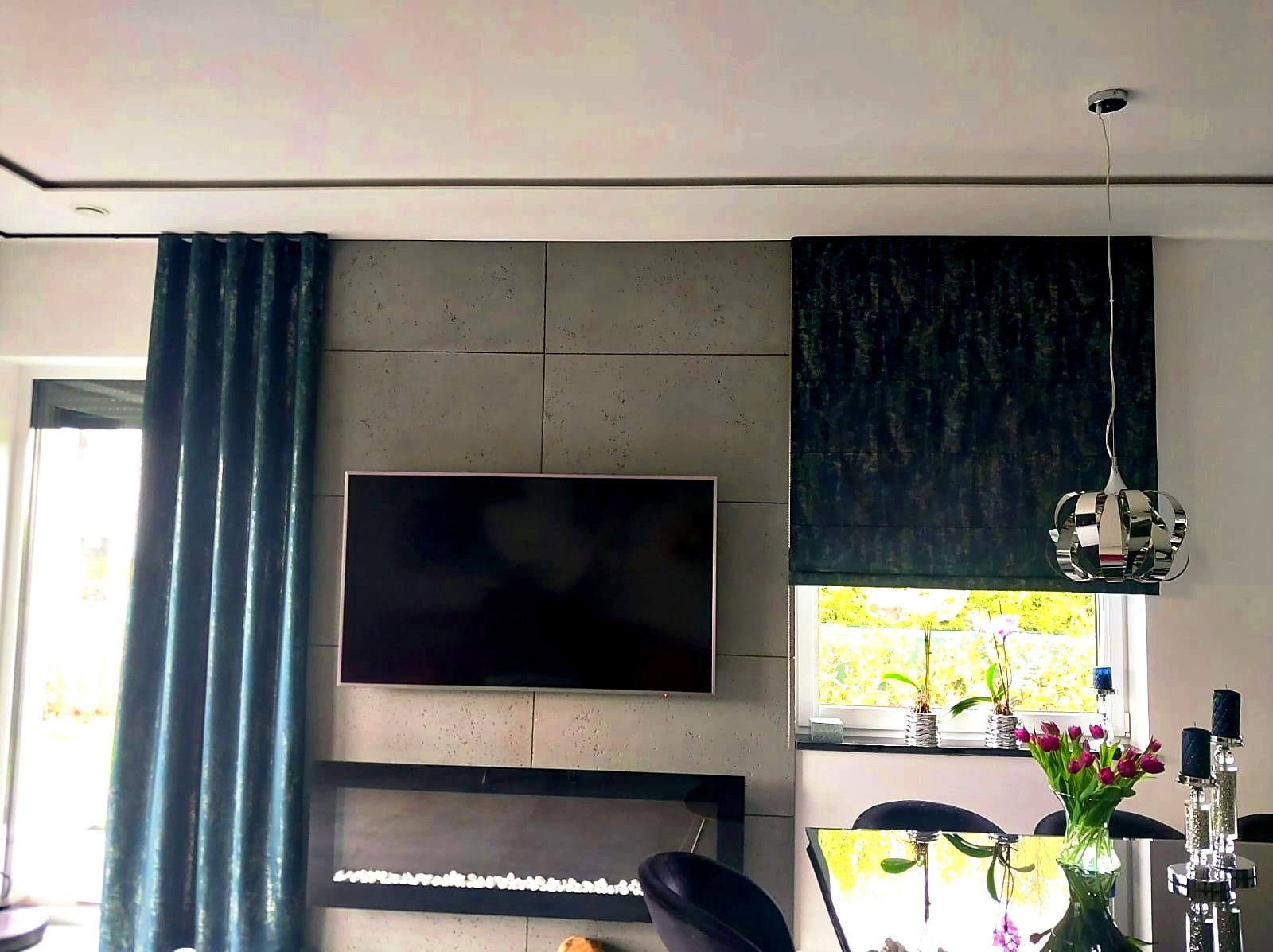 Nowoczesny salon z zasłonami w kolorze morskim i roletą rzymską w odcieniach szarości, telewizor na ścianie z betonowych paneli, pod nim kominek elektryczny, wisząca lampa nad stołem z bukietem...