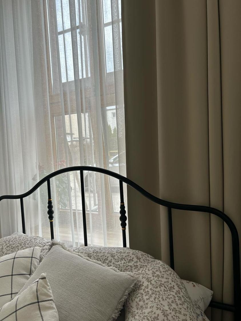 Sypialnia z czarnym metalowym łóżkiem, wzorzystą pościelą i zwiewnymi białymi zasłonami w oknie, obok zasłony w kolorze beżowym.