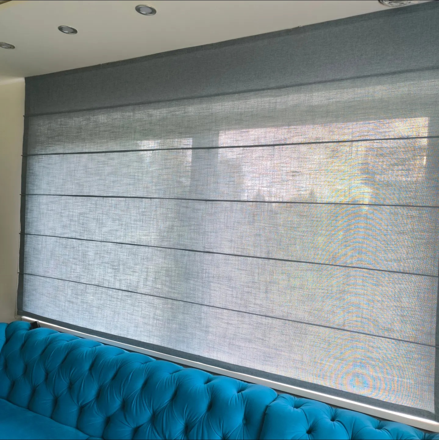 Szara, tekstylna osłona okienna z poziomymi zakładkami, zamontowana nad turkusową sofą z pikowaniem.
