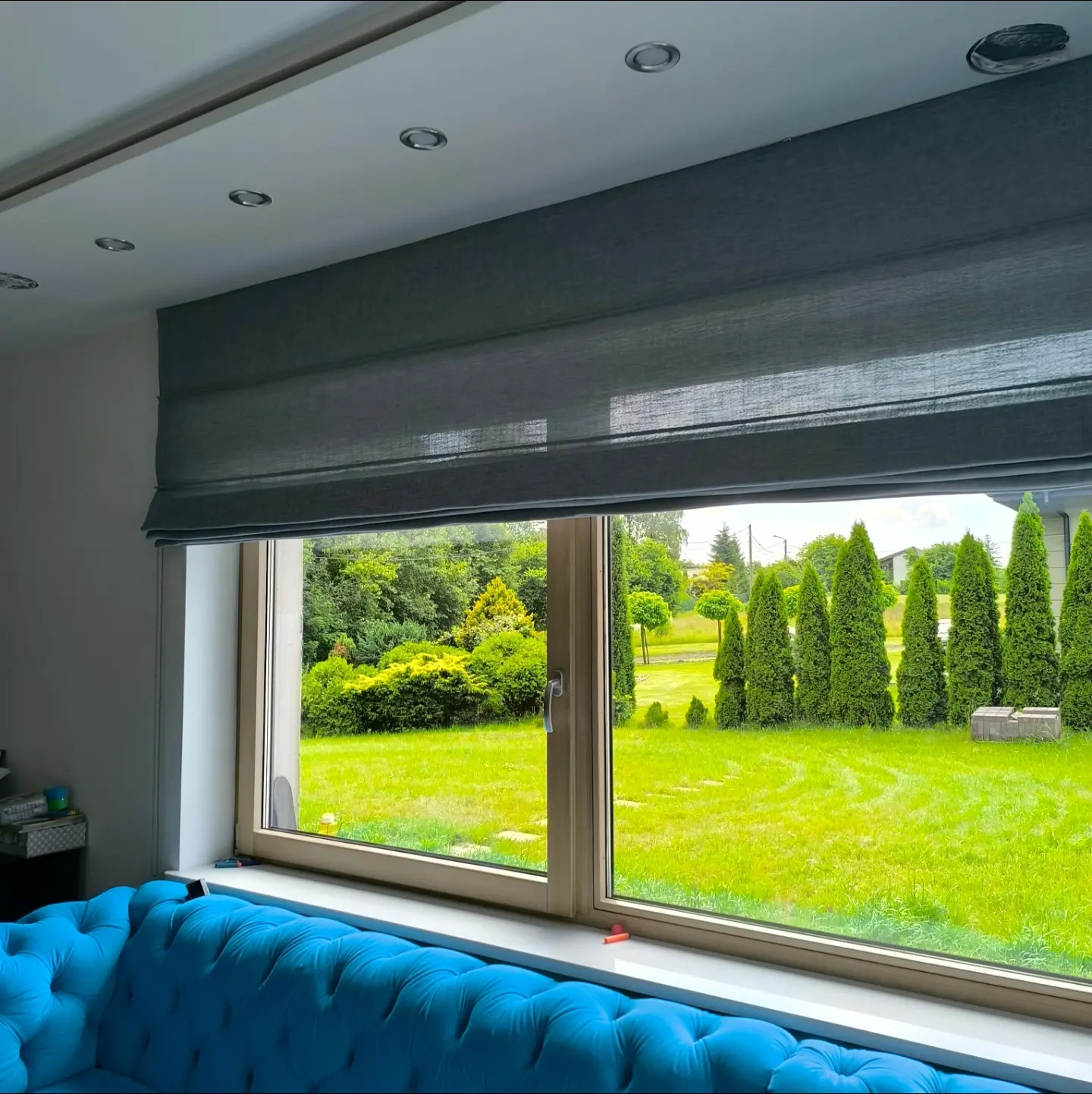 Szara, zwinięta roleta materiałowa w oknie z widokiem na zadbany ogród z tujami i trawnikiem; w tle fragment turkusowej sofy.