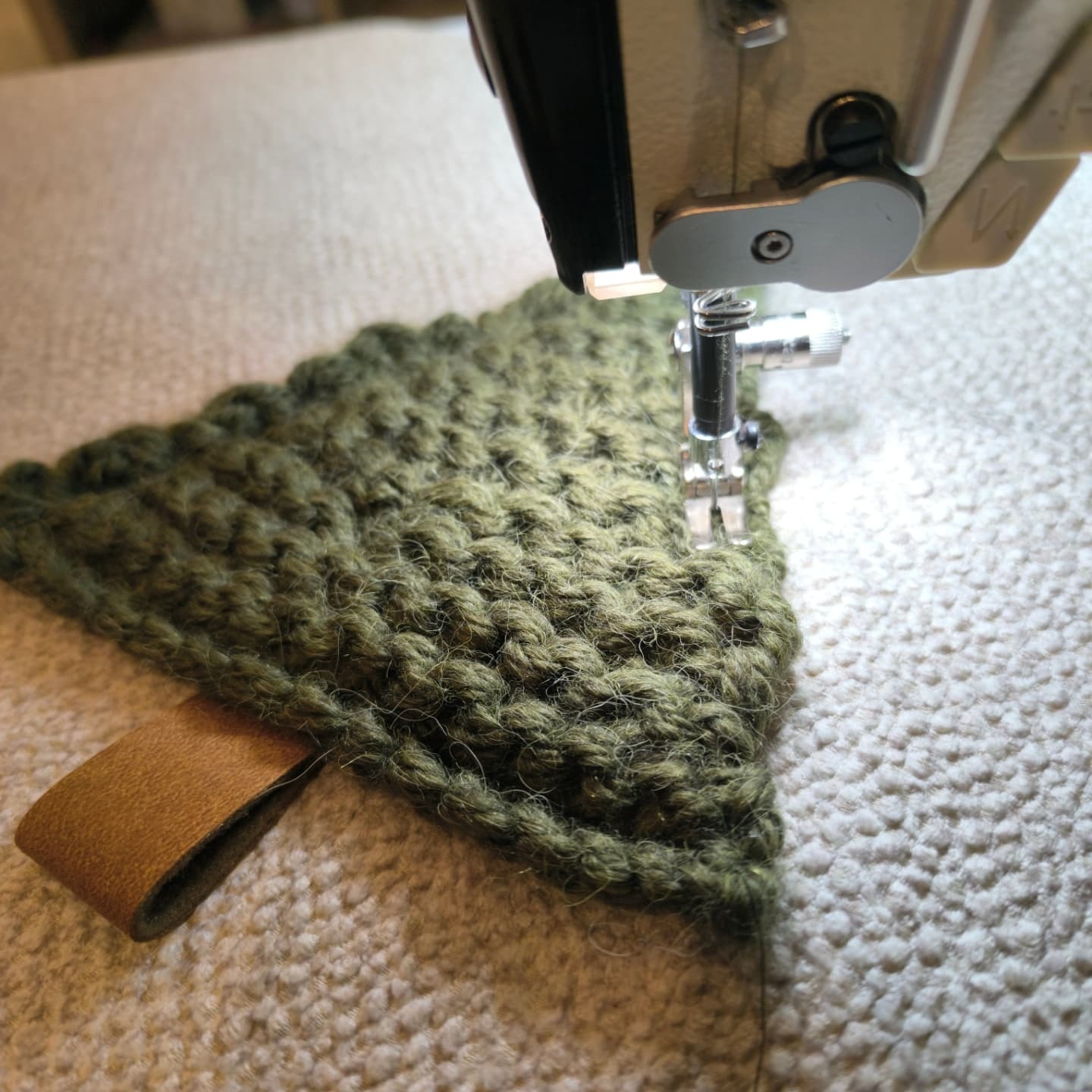 Fragment szycia na maszynie, zielona dzianina o trójkątnym kształcie z brązową skórzaną pętelką przyszywana na beżowej tkaninie.