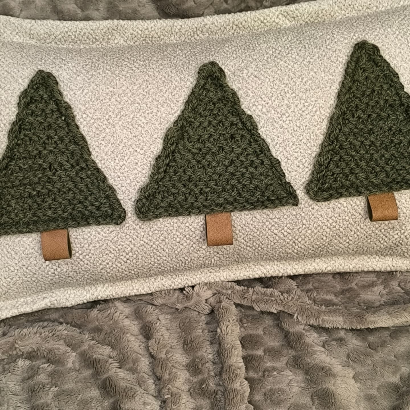 Dekoracyjna poduszka z trzema naszytymi, zielonymi choinkami z włóczki na brązowych pniach, leżąca na szarym, pluszowym kocu.