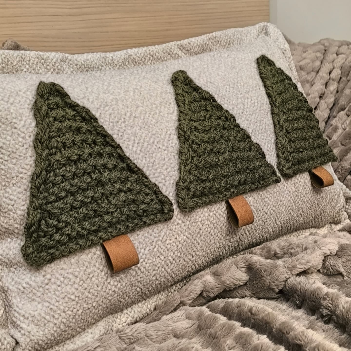 Dekoracyjna poduszka z trzema naszytymi, szydełkowanymi choinkami w odcieniach zieleni, każda z brązową skórzaną zawieszką u podstawy, leżąca na puszystym, szarym kocu.