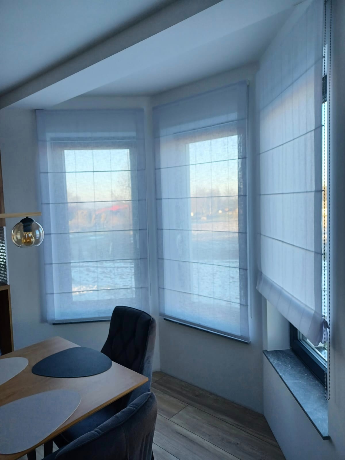 Jasne, półprzezroczyste rolety w stylu rzymskim zamontowane w trzech oknach, widoczne z wnętrza jadalni z drewnianym stołem i szarymi krzesłami.